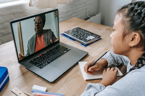 Free From above of focused Asian girl sitting at table with copybook and pencil and carefully listening to black teacher explaining homework task during videocall on laptop Stock Photo